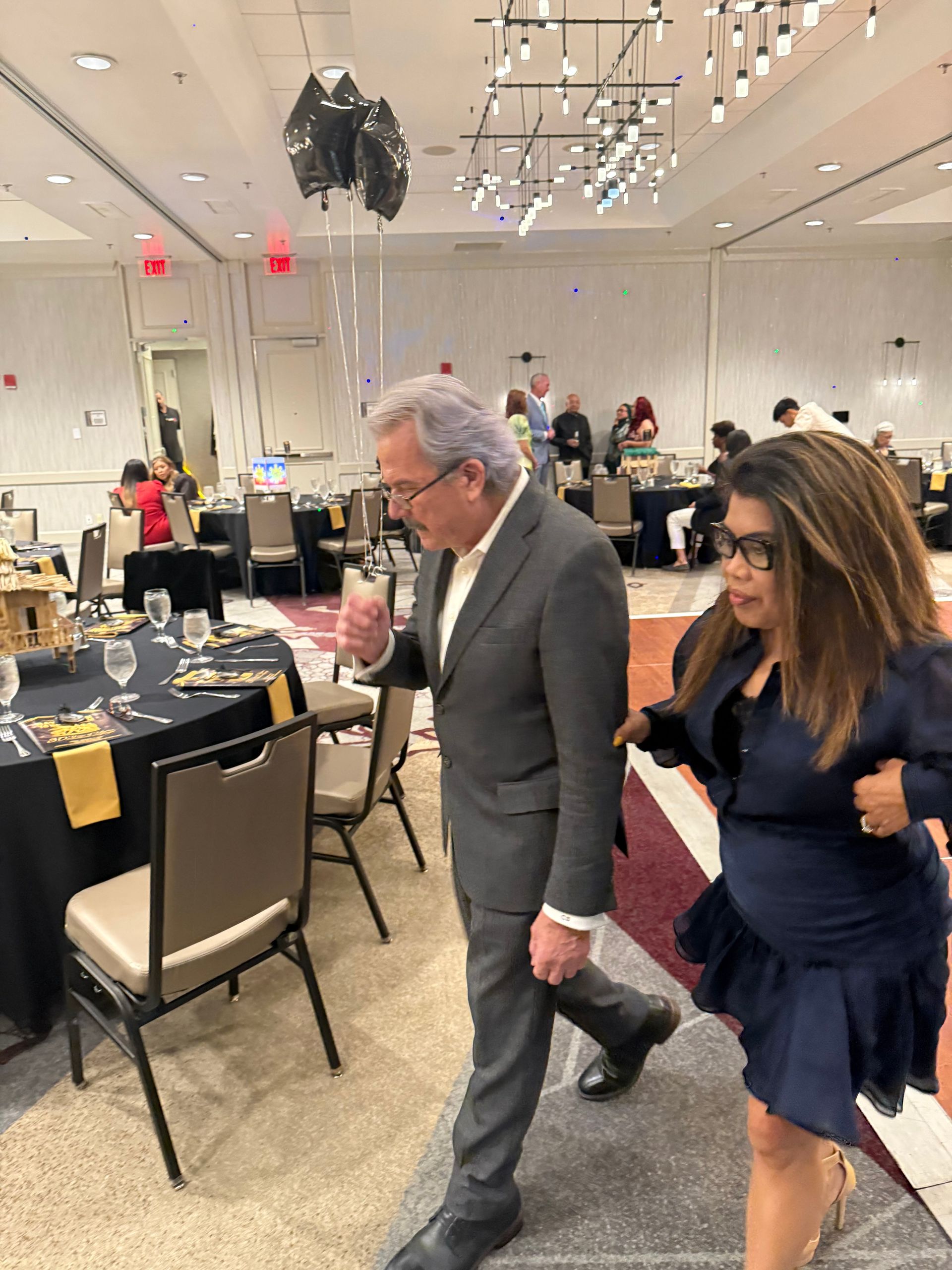 Man in suit and woman in dress walking in event venue. Black table settings, lights.