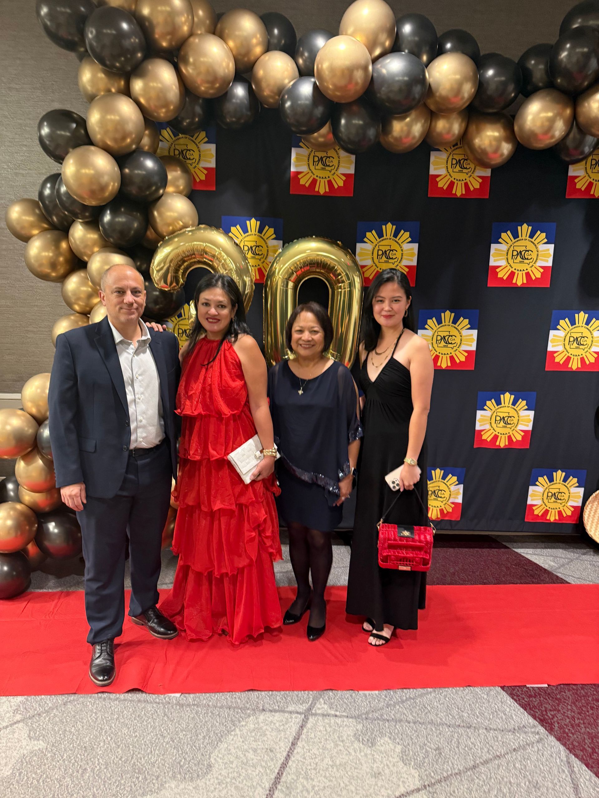 Four people pose on a red carpet in front of a backdrop with gold and black balloons.
