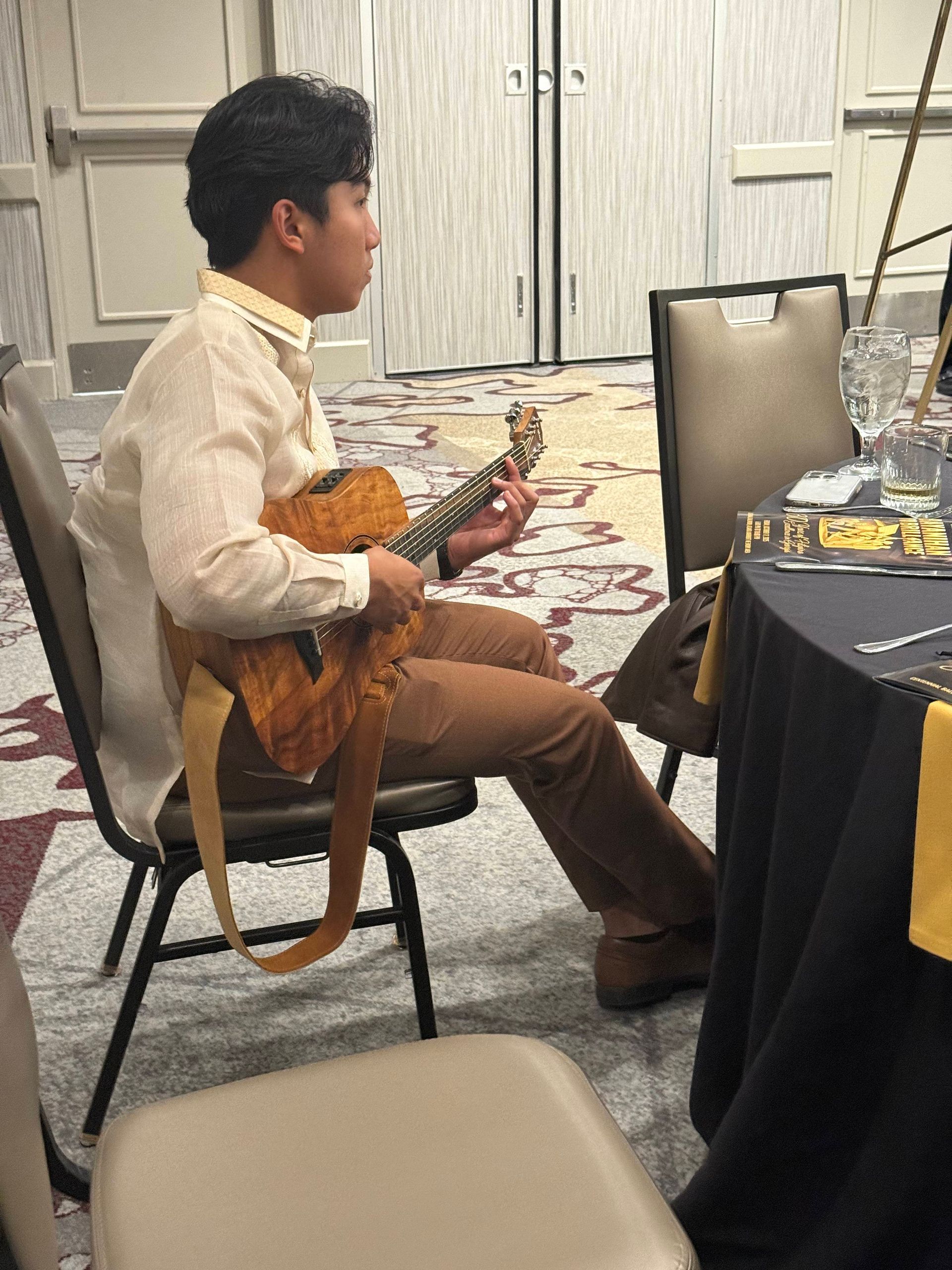 Man playing guitar at a table in a room. He wears a cream shirt and brown pants.