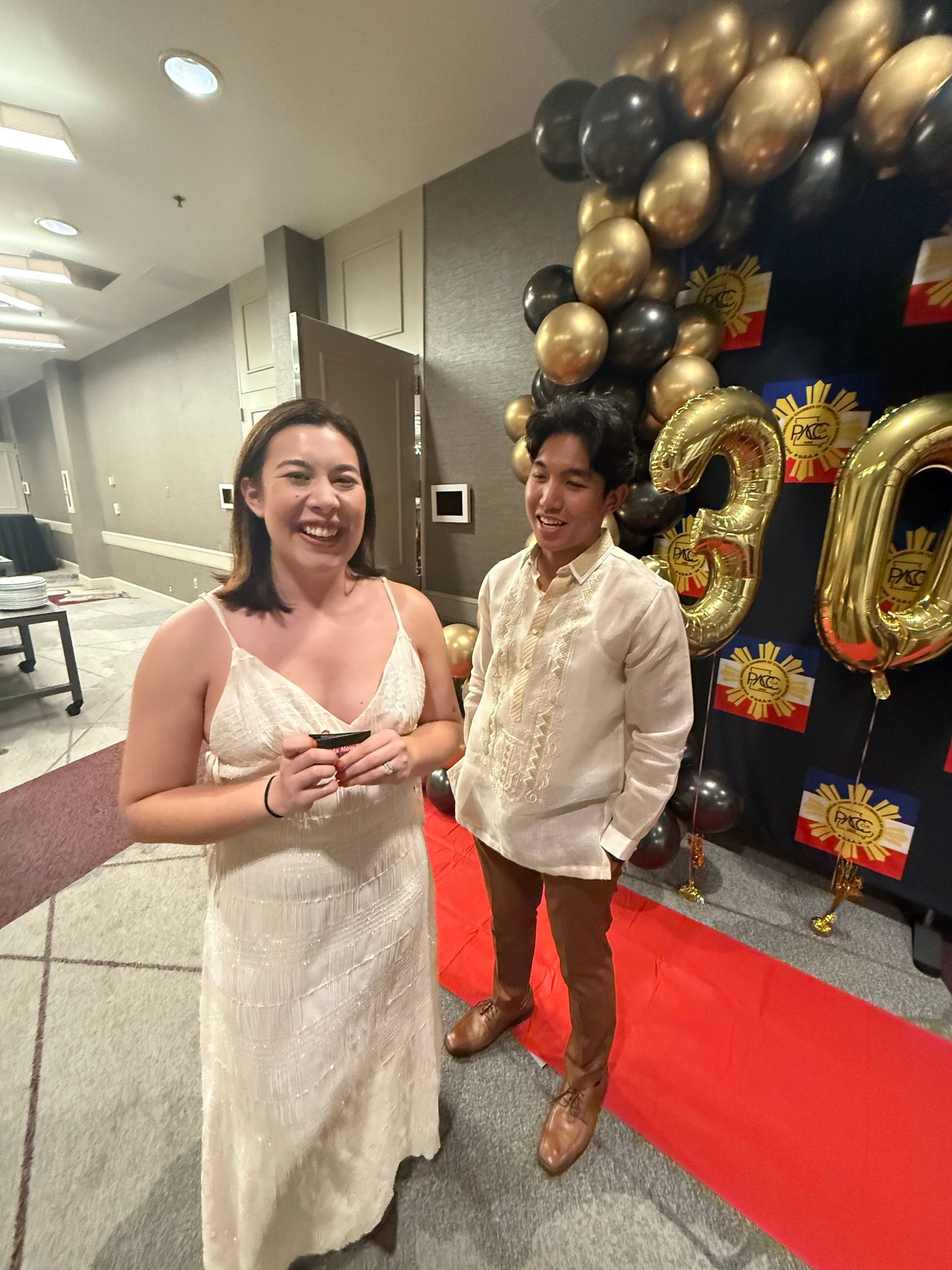 Woman in white dress smiles, man in beige shirt stands near gold and black balloons; formal event.