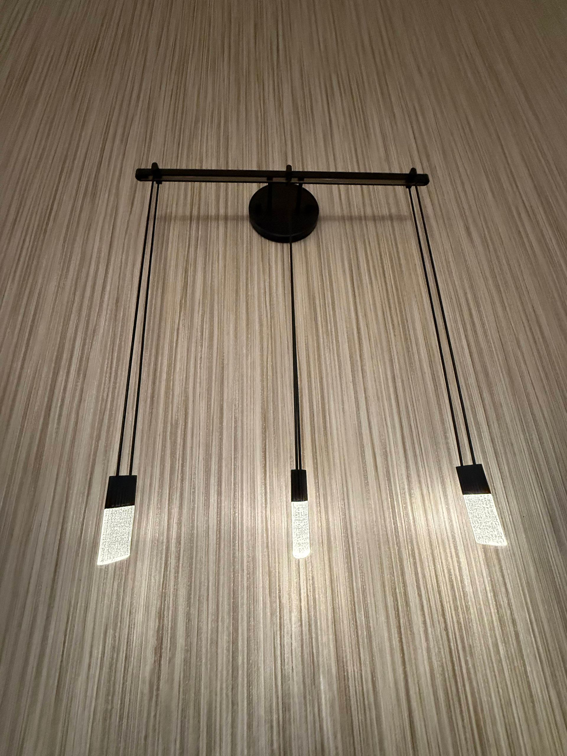 Three-light fixture with black metal rods and clear, rectangular light pendants against a textured wall.