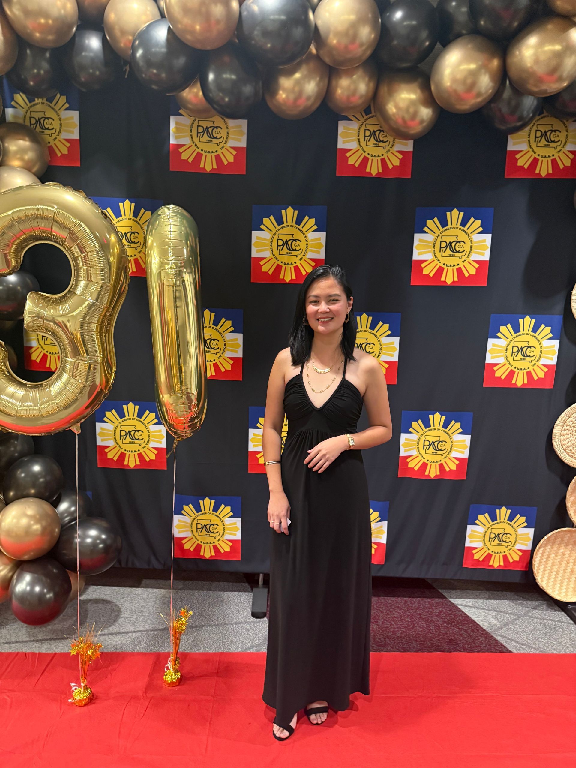 Woman in a black dress stands on a red carpet with balloons and Philippine flags in the background.