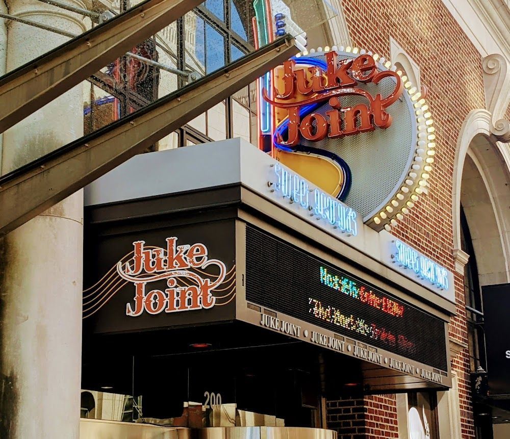 Juke Joint sign on a building with brick facade. Neon lights and a digital marquee display show the venue’s name and event information.