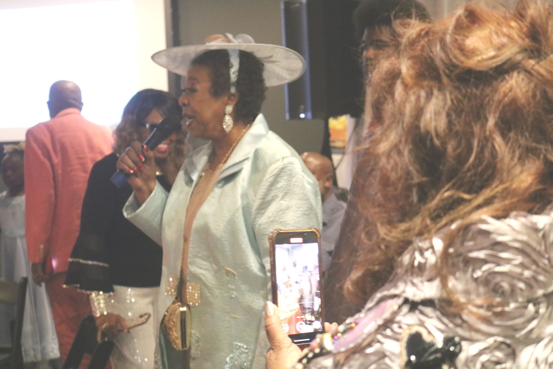 Woman in light blue jacket and hat speaks into microphone. Others in the background.