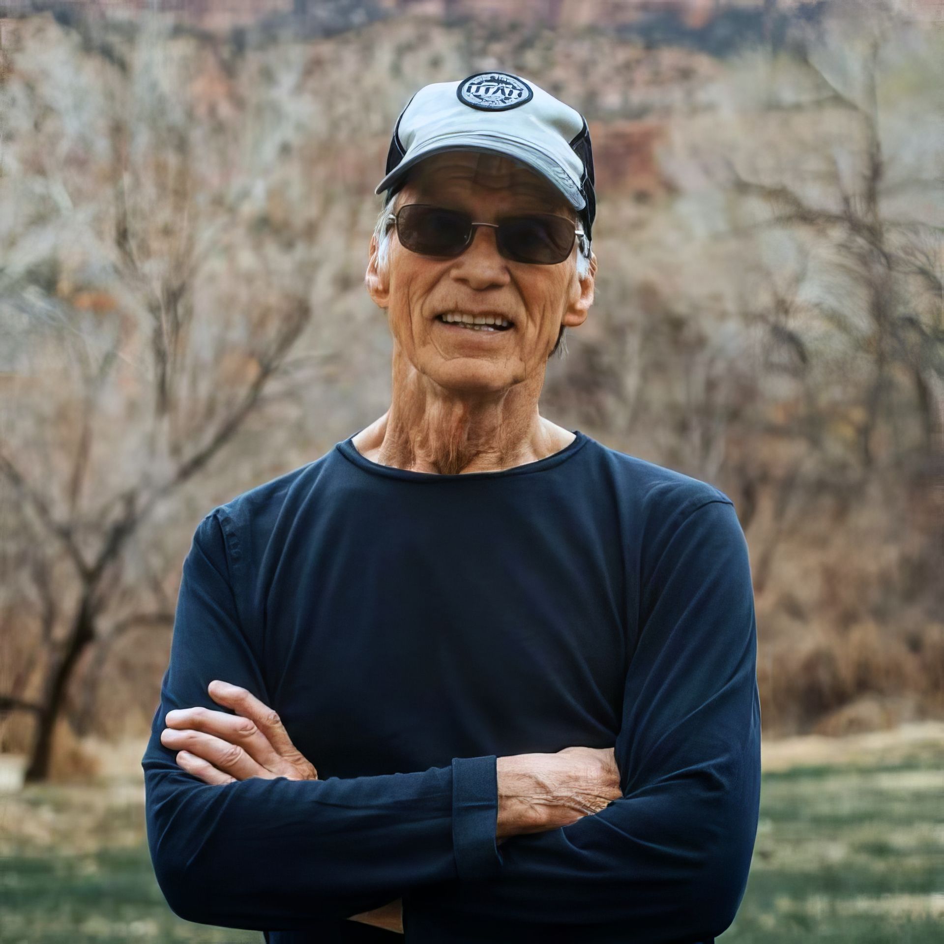Man in hat and sunglasses with arms crossed, standing outdoors in front of a tree-lined backdrop.