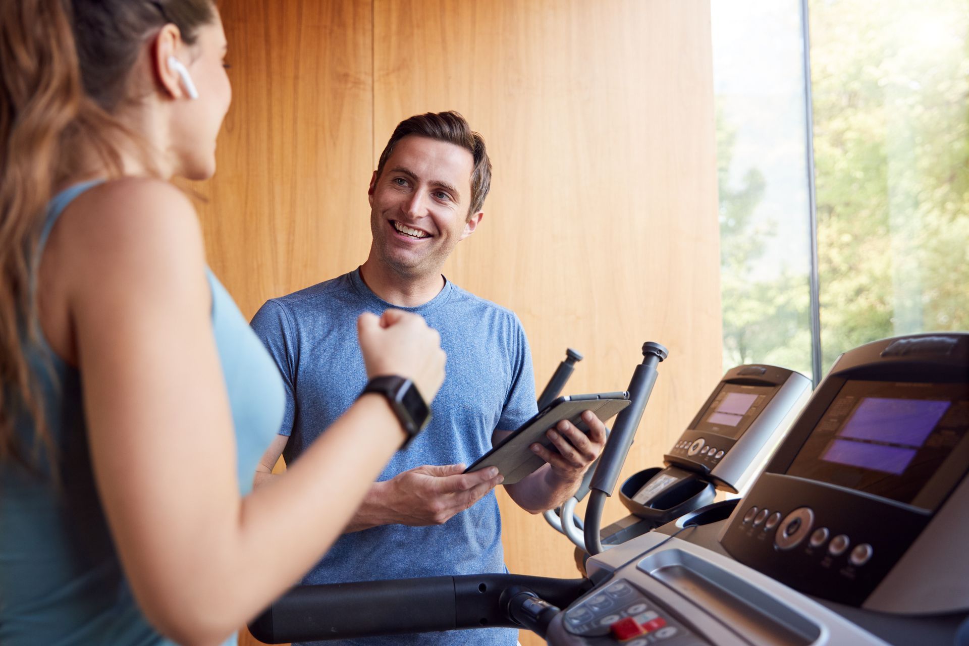 A trainer smiles at a woman on a treadmill. They're in a gym with wooden panels and a large window.