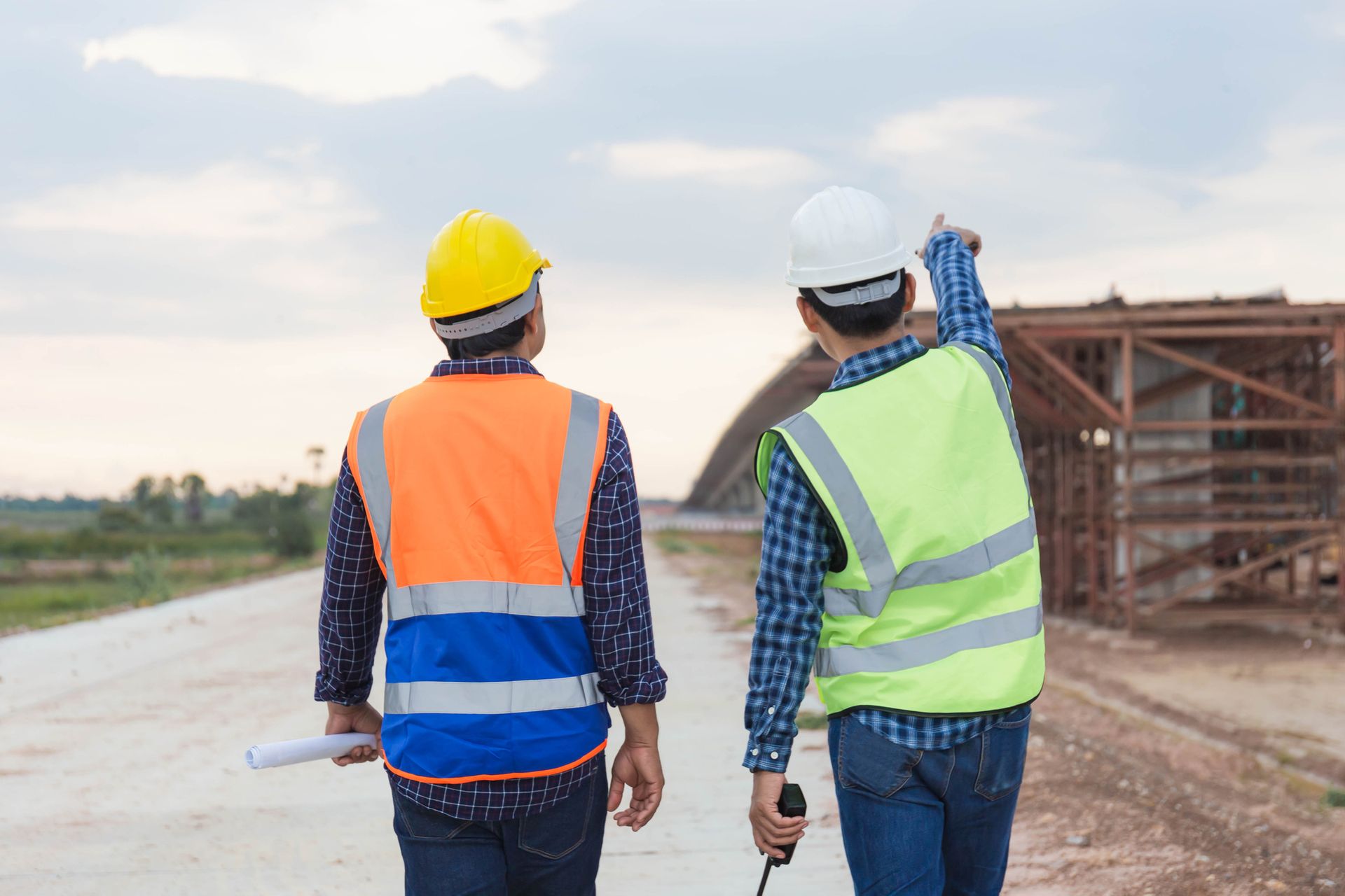 Two construction workers in safety vests and hard hats on a construction site, one pointing.