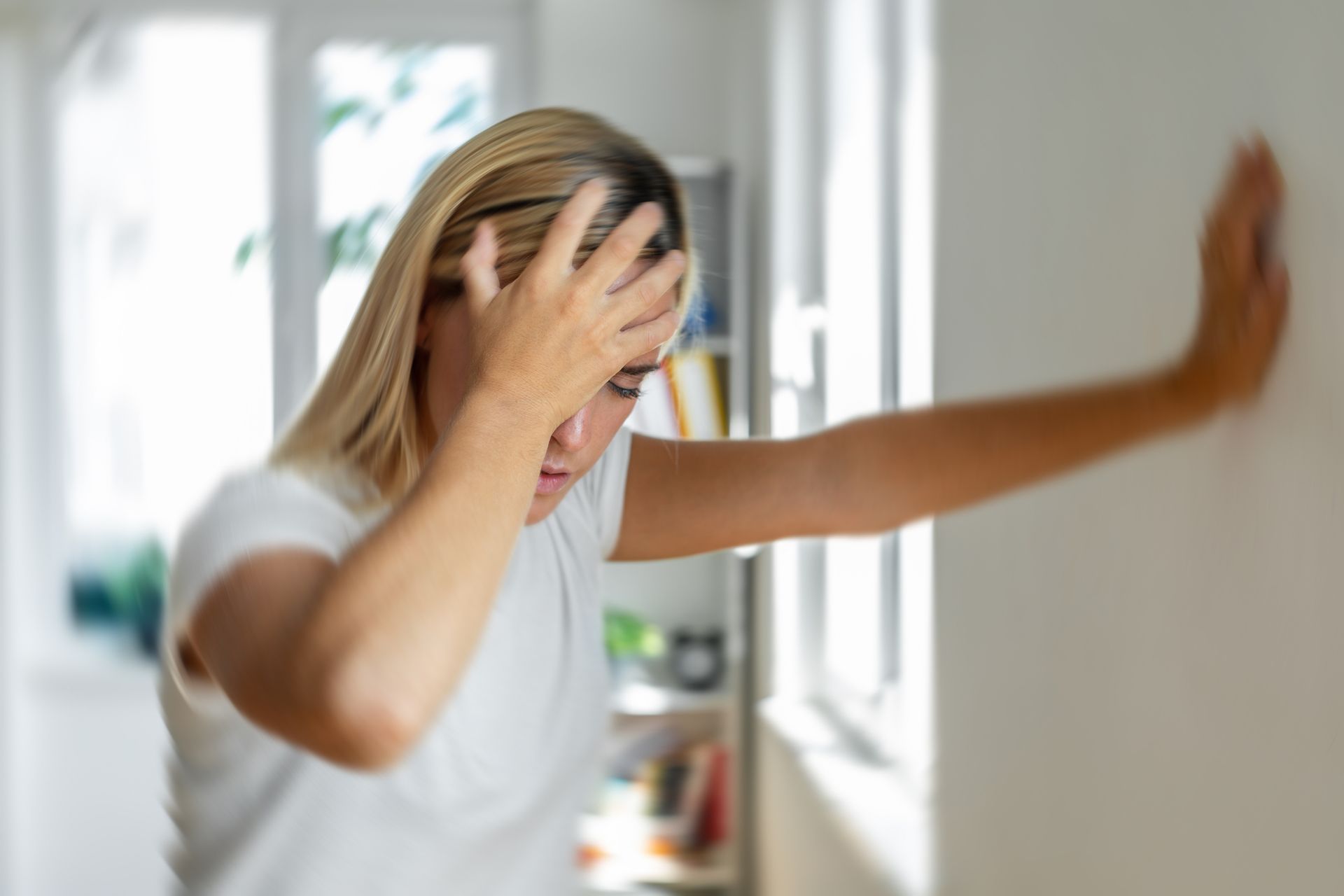 Woman with blond hair, holding her head and leaning against a wall, appearing dizzy in a bright room.