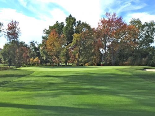 Green golf course with trees showing fall colors against a partly cloudy sky.