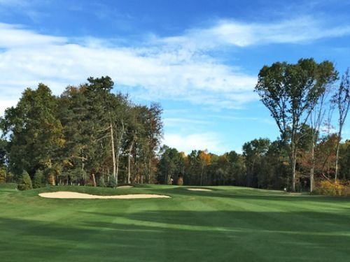 Green golf course with sand traps and trees under a partly cloudy blue sky.