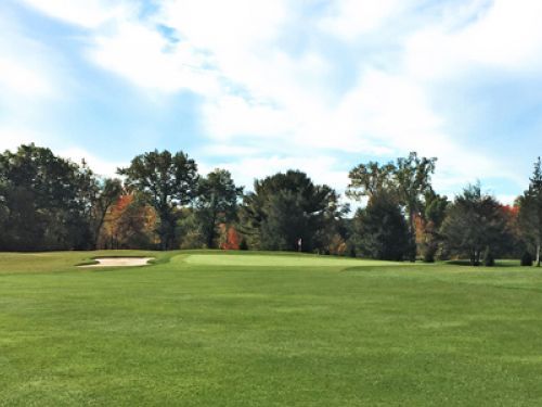 Golf course with green grass, trees, and a sand trap under a blue sky.
