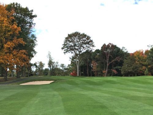 Green golf course with a sand trap, trees with fall foliage, and an overcast sky.