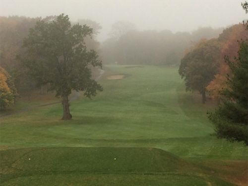 Golf course fairway on a foggy day; trees line the edges, green grass, and sand trap visible in the distance.