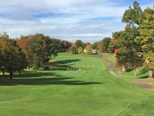 Green golf course with trees changing colors under a partly cloudy sky.