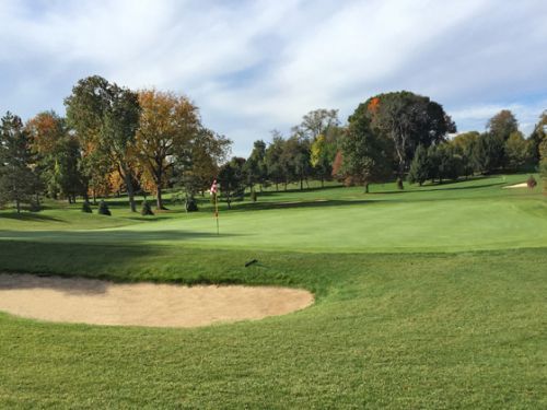 Green golf course with a sand trap, flag on the green, and fall foliage in background.
