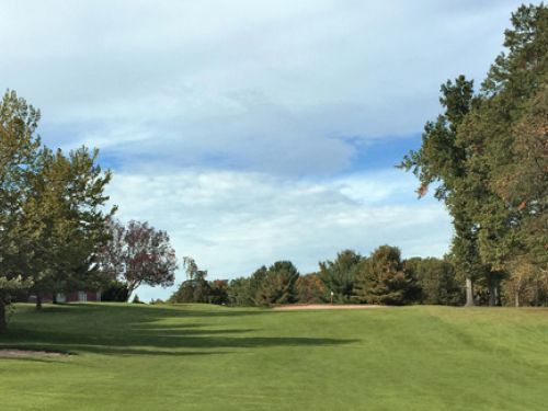 Green golf course under a partly cloudy blue sky, with trees on either side.