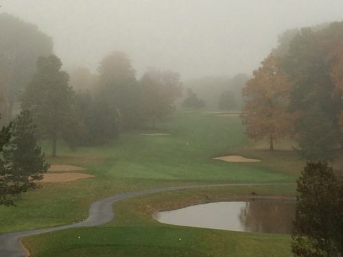 Golf course in heavy fog; green fairway, pond, and trees visible.