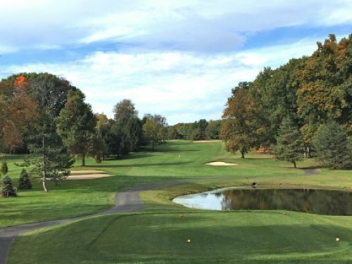 Golf course with green fairway, water hazard, trees, and blue sky.