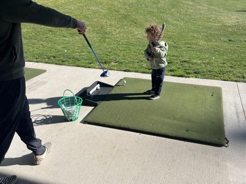 Young child at driving range, holding golf club, being coached by adult.