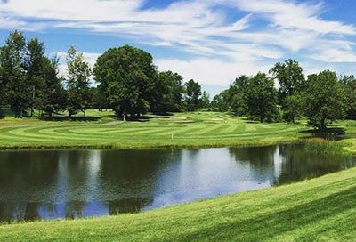 Golf course with green grass, trees, and a lake under a blue sky with clouds.