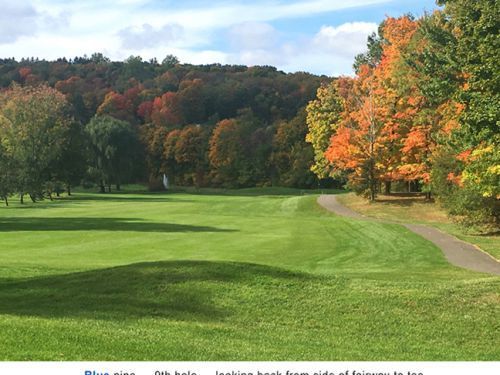 Golf course with green fairway, fall foliage in orange and red, trees in the background, blue sky.
