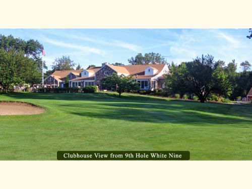 Clubhouse view from golf course. Green lawn, trees, blue sky, and a large building with a brown roof.