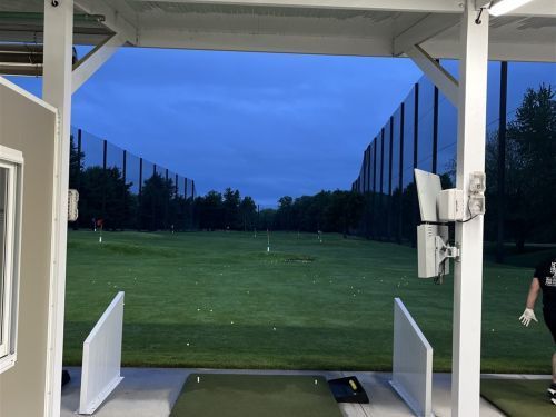 View from a golf driving range stall; green field with targets and protective netting. A person is visible on the right.