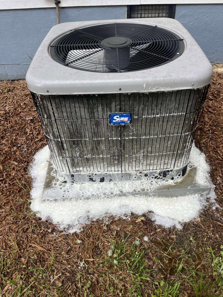 An air conditioning unit with foam around the base, likely from cleaning. Outdoors, in a yard.