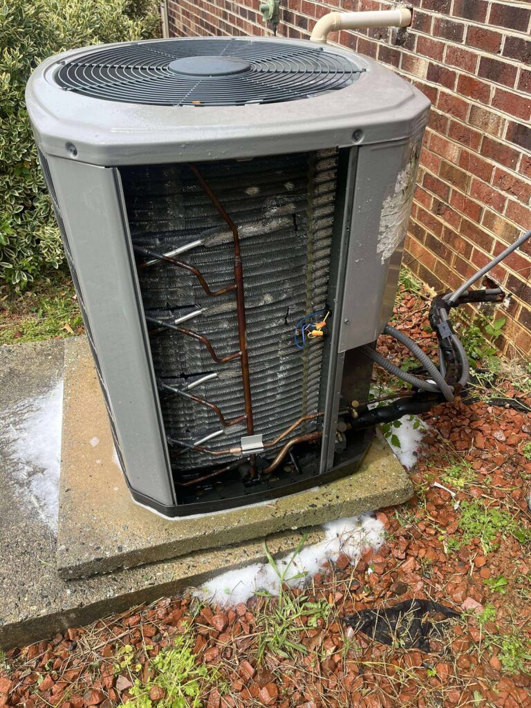 An air conditioning unit with open side panel on a concrete slab near a brick wall and vegetation.
