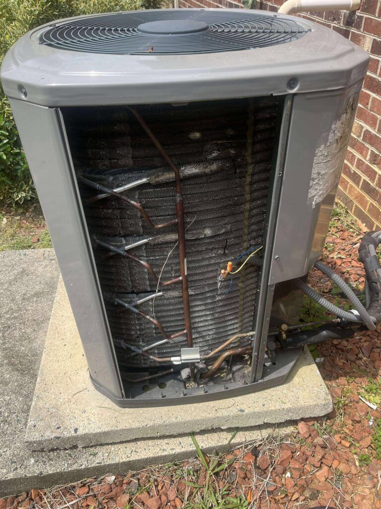 AC unit with open panel, showing dirty coils, sitting on concrete pad next to brick wall.