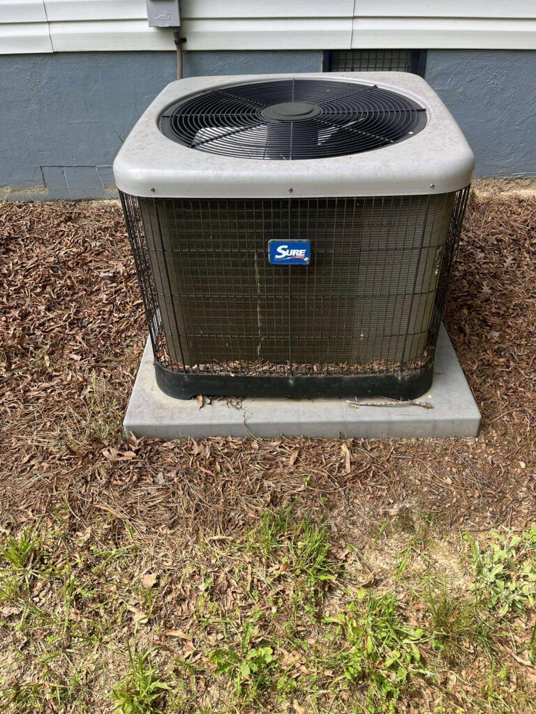 Air conditioner unit, gray and black, sitting on concrete base near a building's foundation and grass.