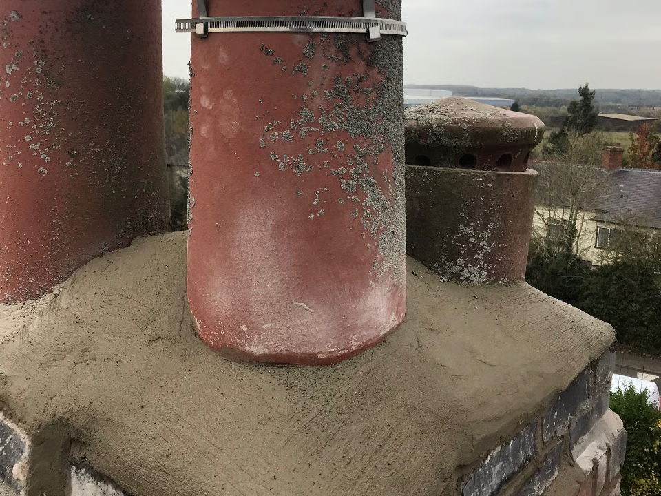 Three red chimney pots on a gray mortar chimney top; overcast sky and houses in the background.