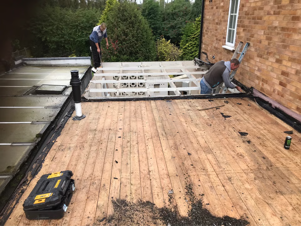 Two people working on a flat roof with wooden beams, near a brick building and greenery.
