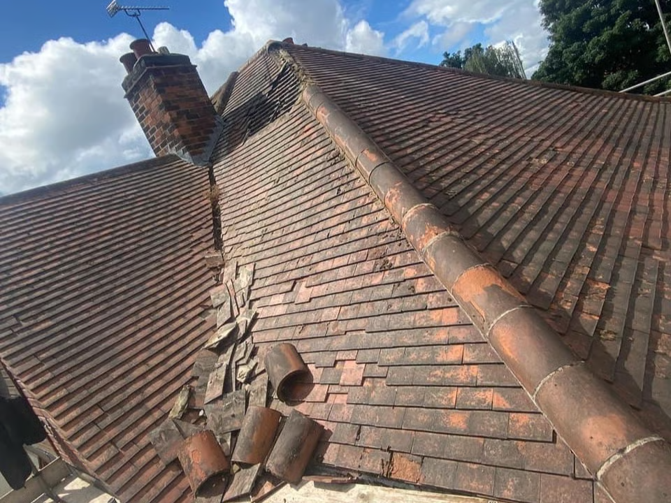 Damaged tiled roof with broken tiles and a chimney under a cloudy sky.