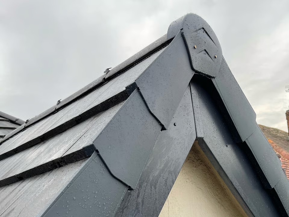 Close-up of a dark grey tiled roof corner with a decorative trim, against a cloudy sky.
