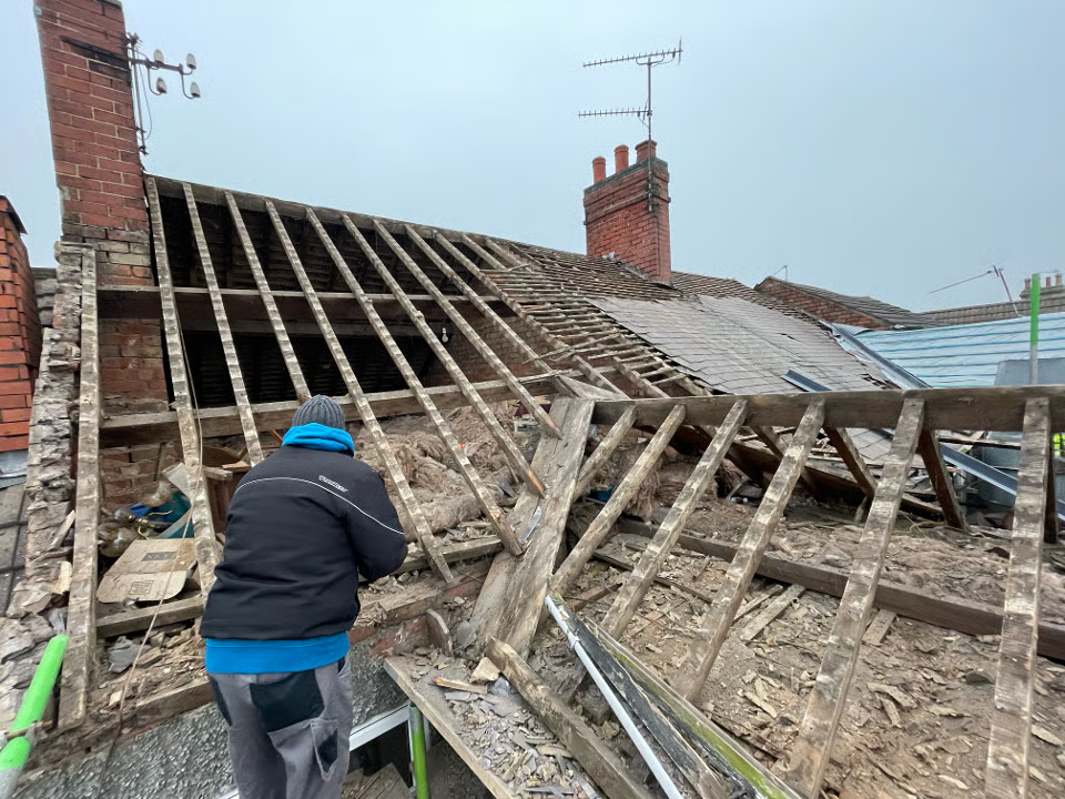 Roofer working on a partially demolished roof, wooden beams exposed. Brick chimney and overcast sky visible.