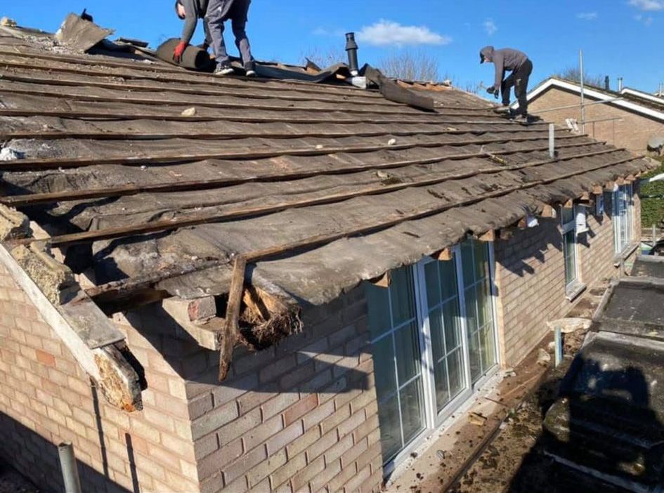 Workers removing old roof tiles from a brick building on a sunny day.