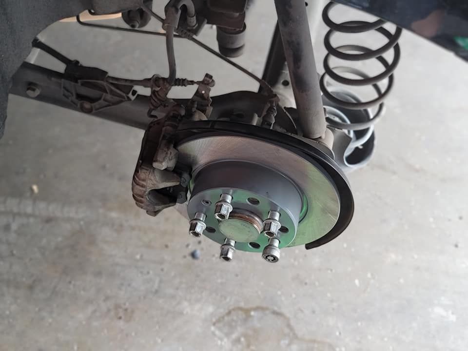 Car's rear wheel with brake rotor and caliper visible. A coil spring and other suspension parts are present.