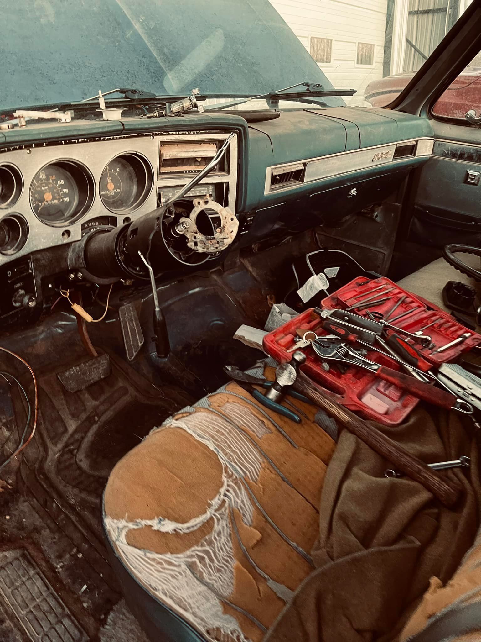 Interior of a teal truck with missing steering wheel. Tools and torn seat visible.