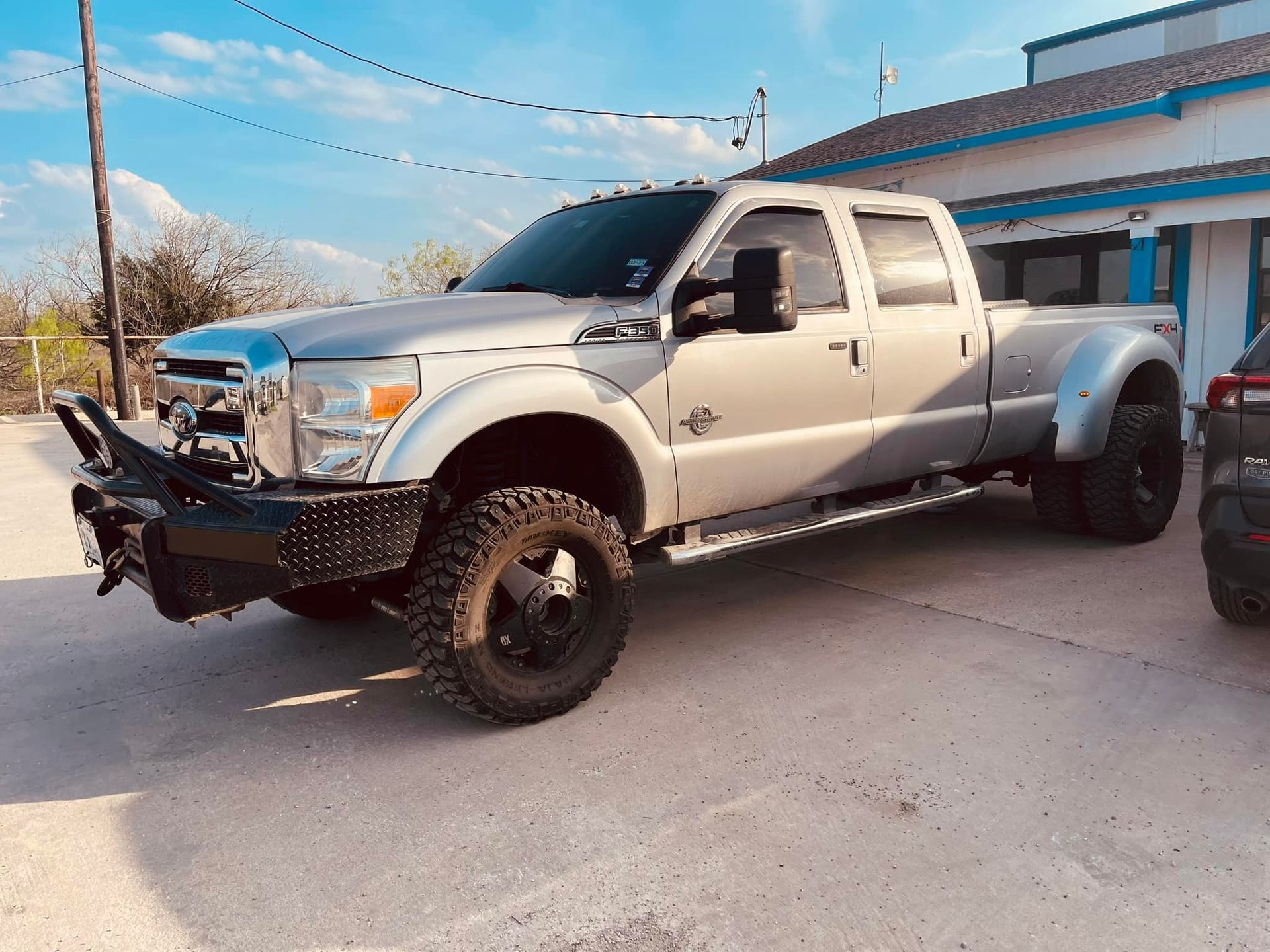 Silver Ford dually pickup truck with a black bumper parked outside a light blue building on a sunny day.
