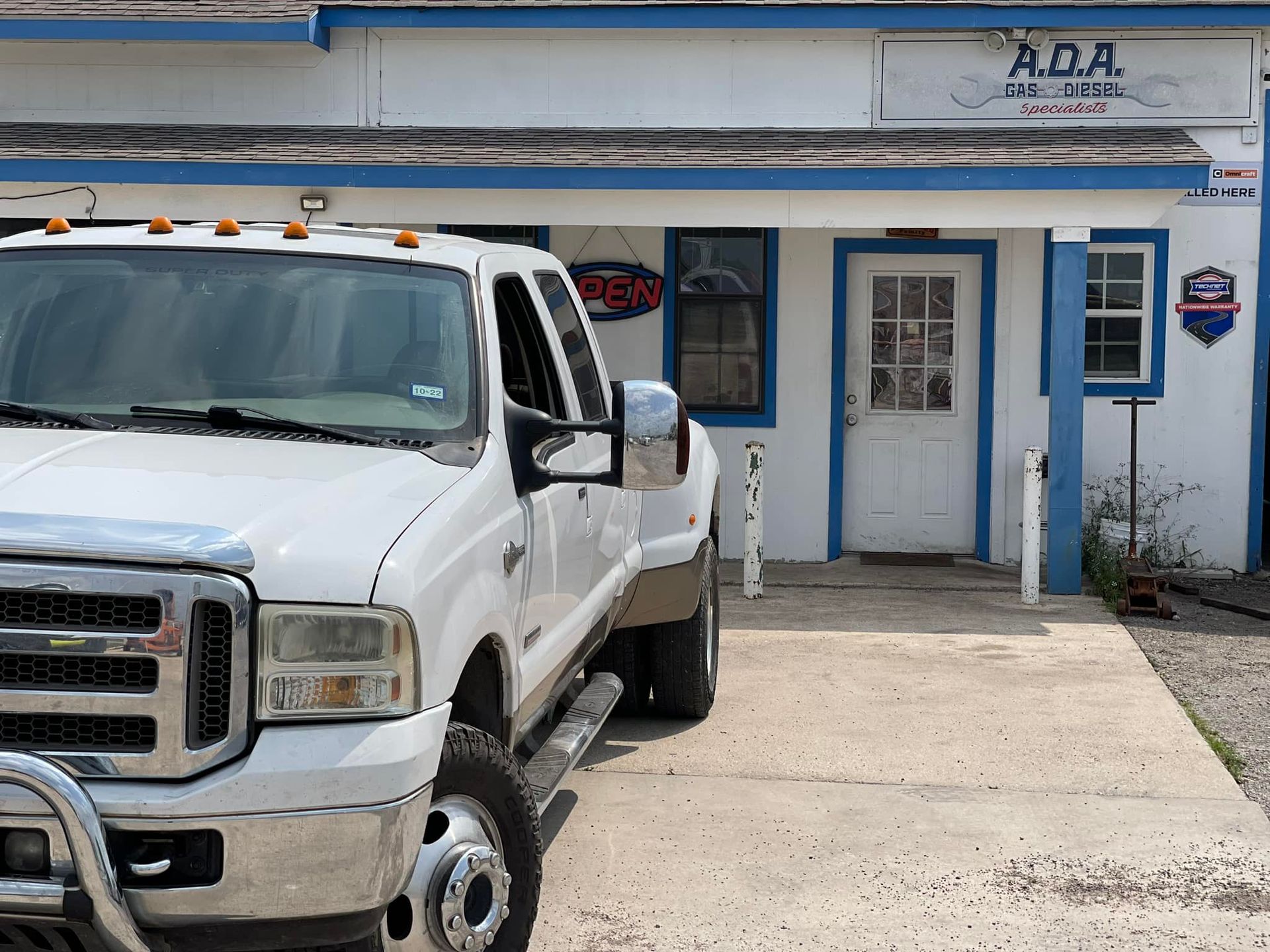 White pickup truck parked in front of a white building with a blue trim and a sign that reads