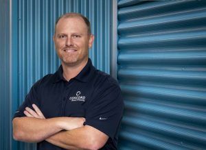 A man in a blue shirt is standing with his arms crossed in front of a blue wall.