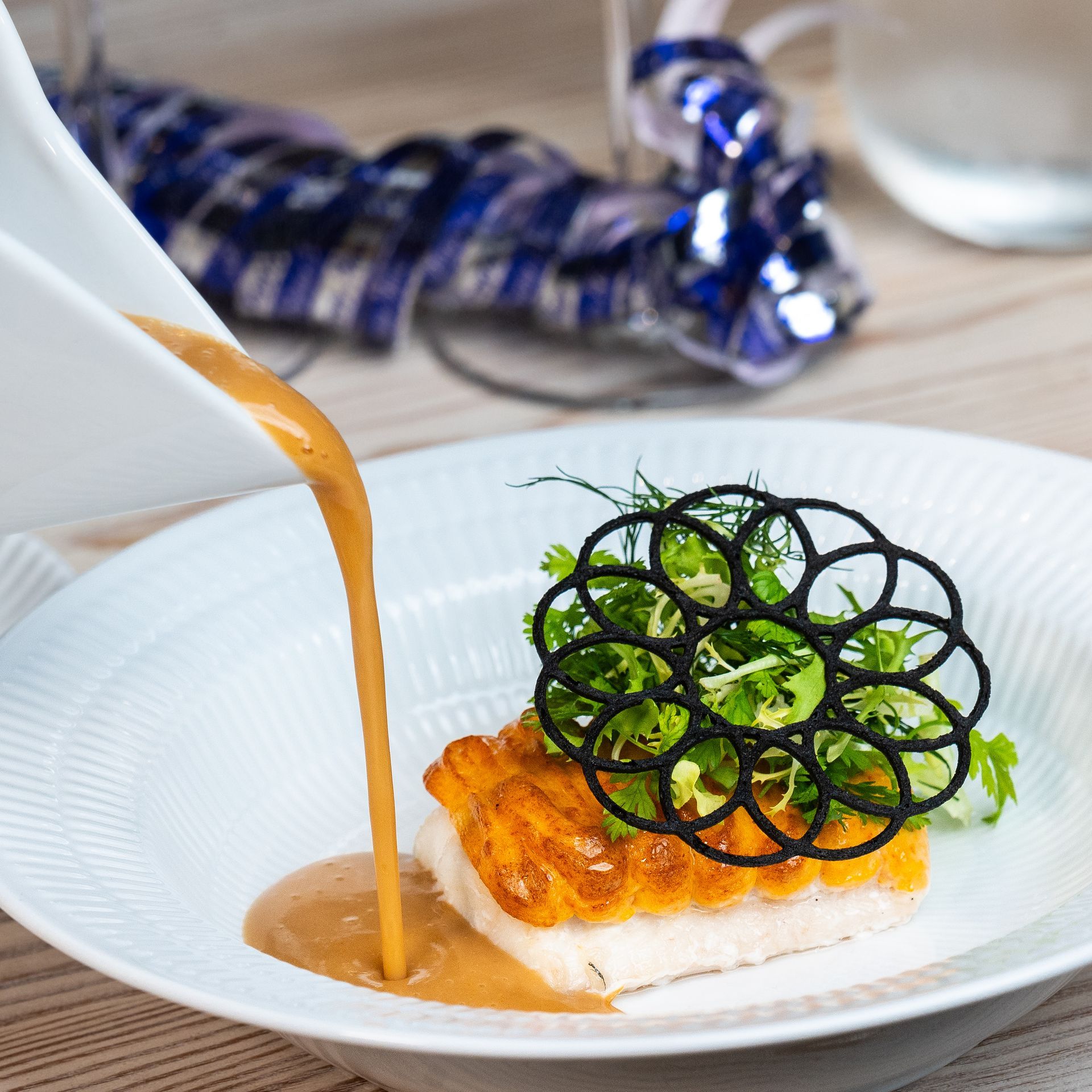 Sauce hældes på en tallerken med fisk, grøntsager og sort dekorativ pynt.