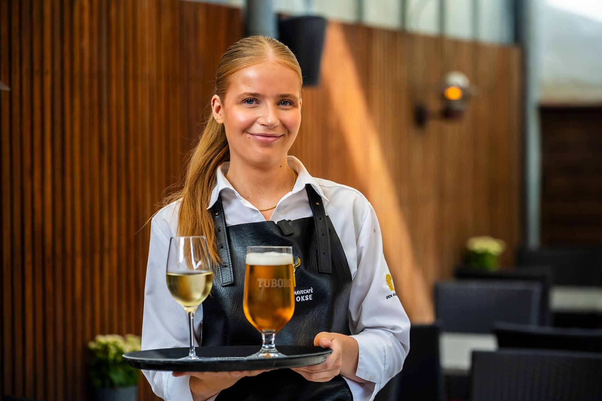 Servitrice smiler og bærer en bakke med hvidvin og øl på en restaurant.
