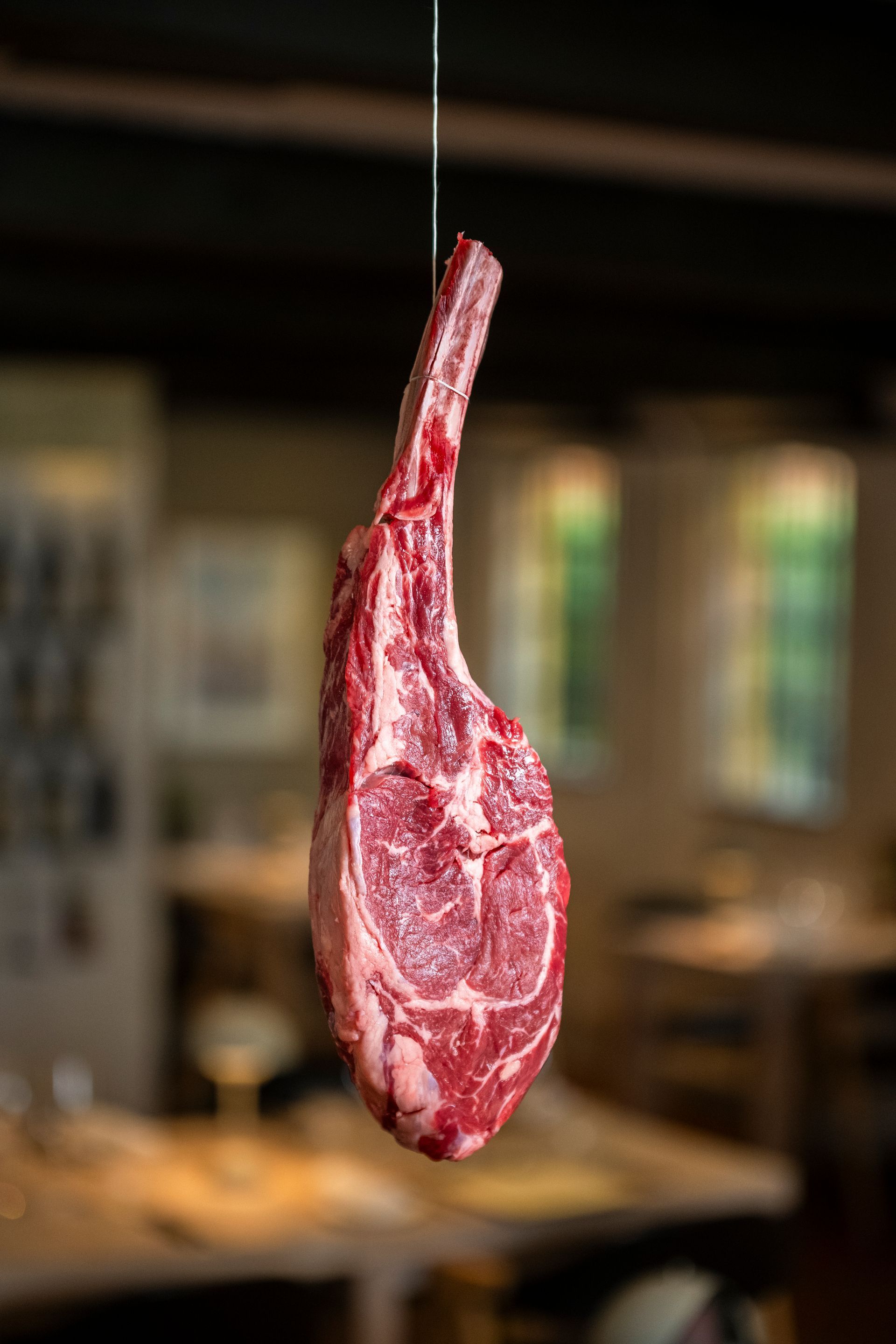 Rå tomahawk-bøf ophængt i en snor i en sløret restaurantomgivelse.
