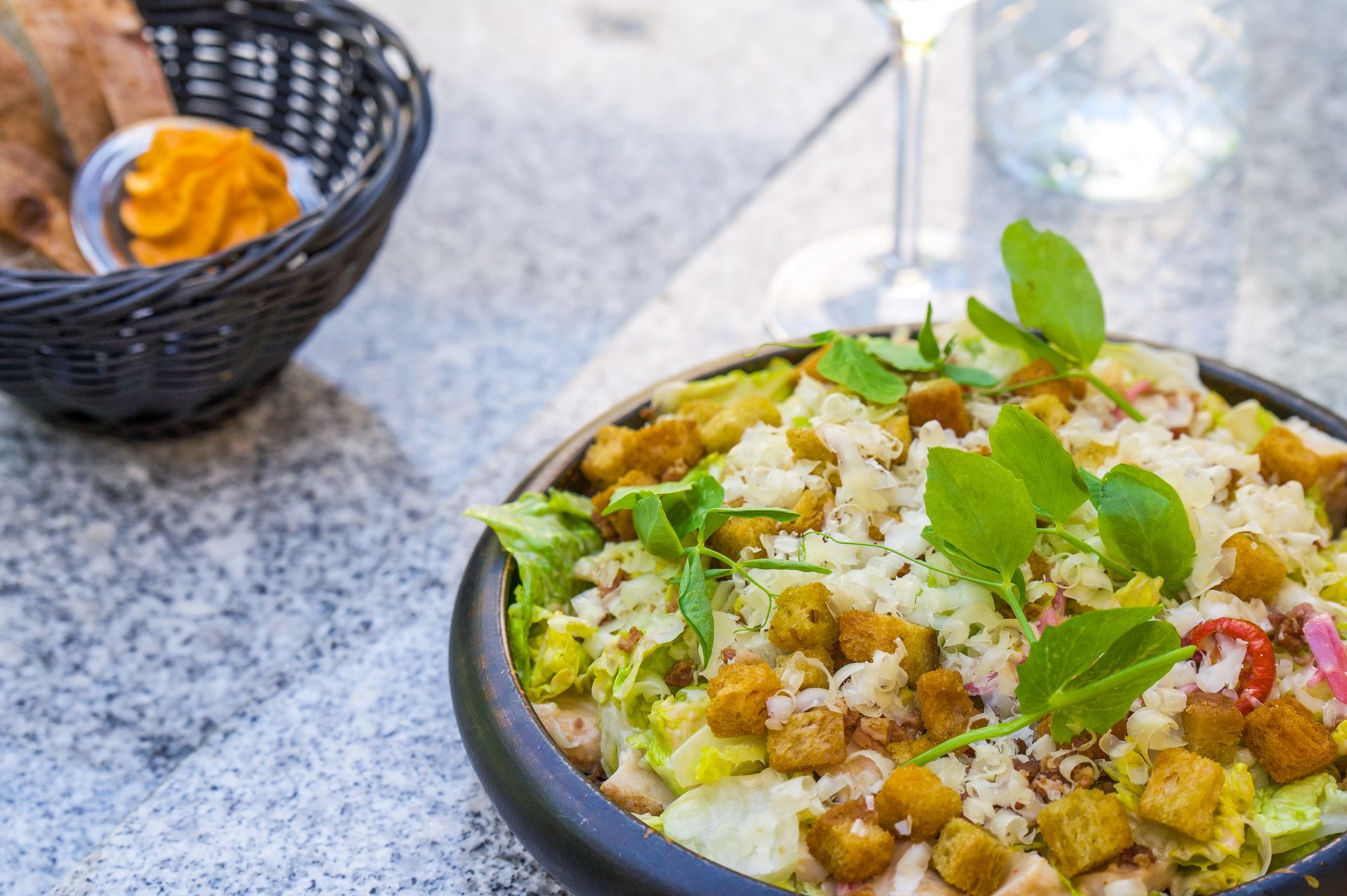 Salat i en sort skål med krutoner, krydderurter og ost på et gråt bord, kurv med brød og dip i baggrunden.