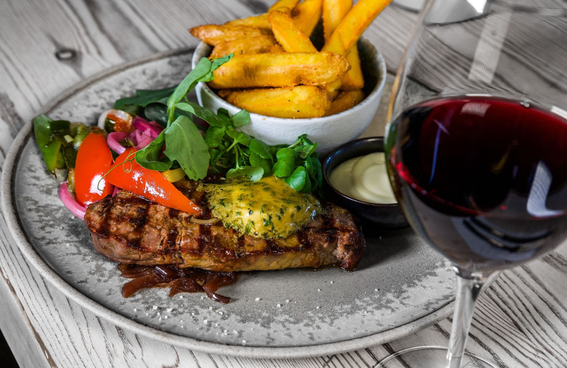 Grillet bøf med pommes frites, grøntsager og vinglas på et træbord.