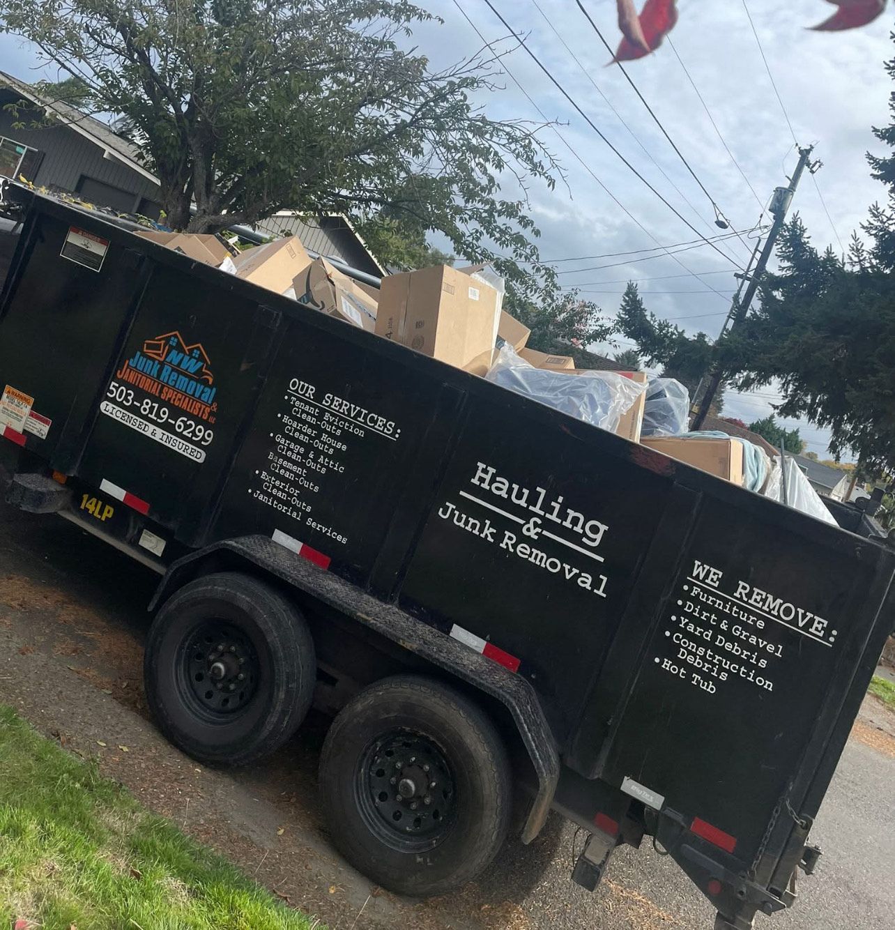 Black junk removal trailer filled with cardboard boxes, parked on a residential street.