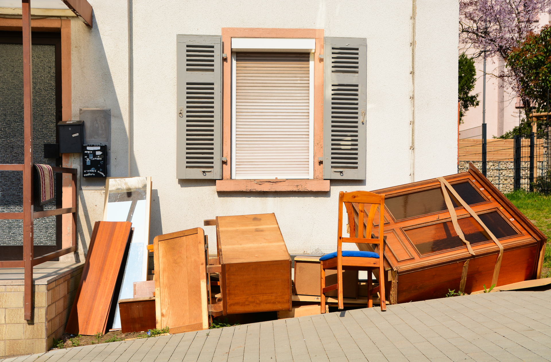 Assorted discarded wooden furniture and items piled outside a white building with an open window.