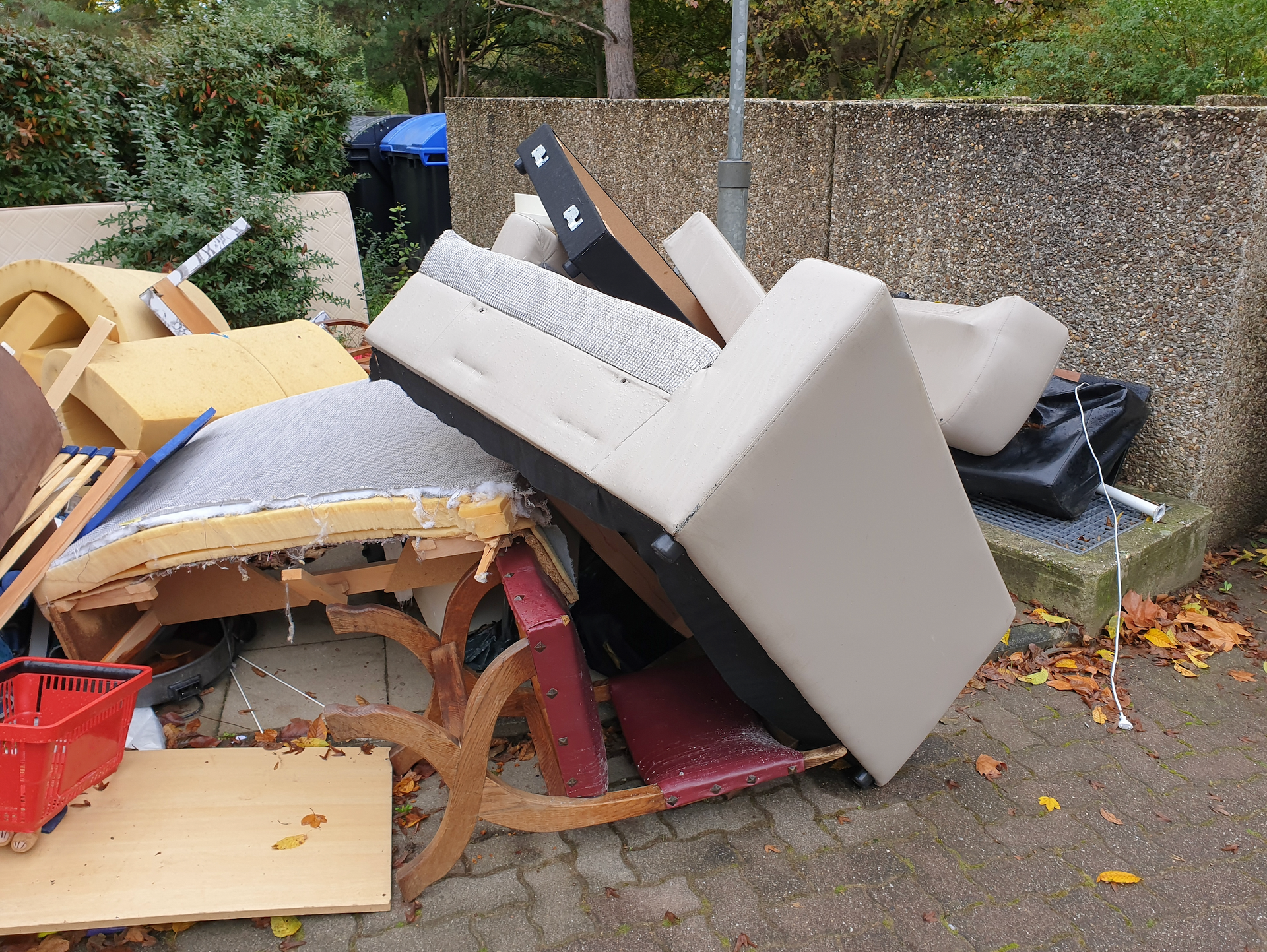 Pile of discarded furniture and debris on a sidewalk next to a concrete wall and a trash bin.
